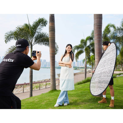 A Godox W-RFT130 130cm (52") Windproof Silver Collapsible Reflector being held by an assistant while a photographer takes photos of a model in a park