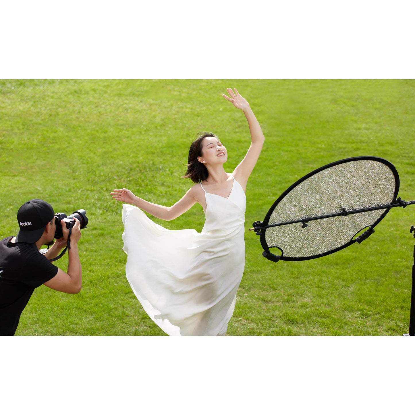 A photographer using a Godox W-RFT80 80cm Collapsible Windproof Reflector on a stand to illuminate a models in a dress in a field