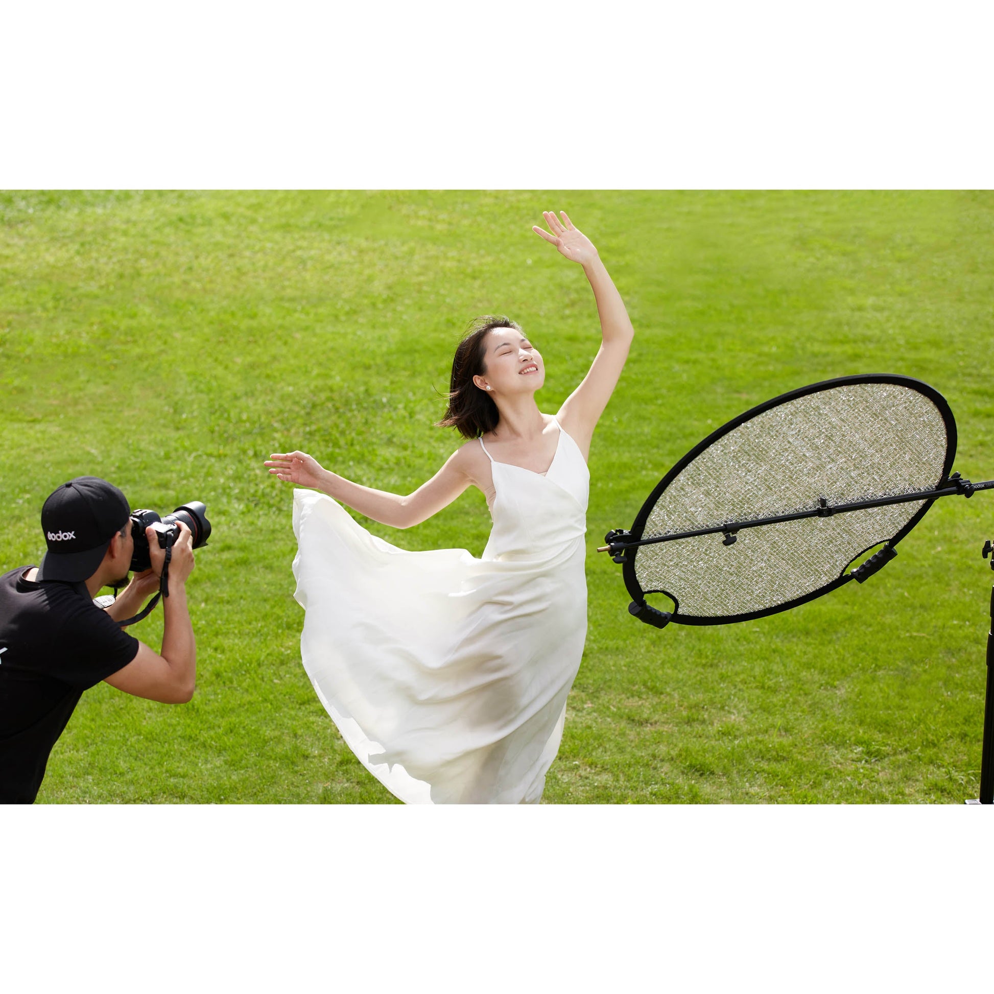 A photographer using a Godox W-RFT80 80cm Collapsible Windproof Reflector on a stand to illuminate a models in a dress in a field