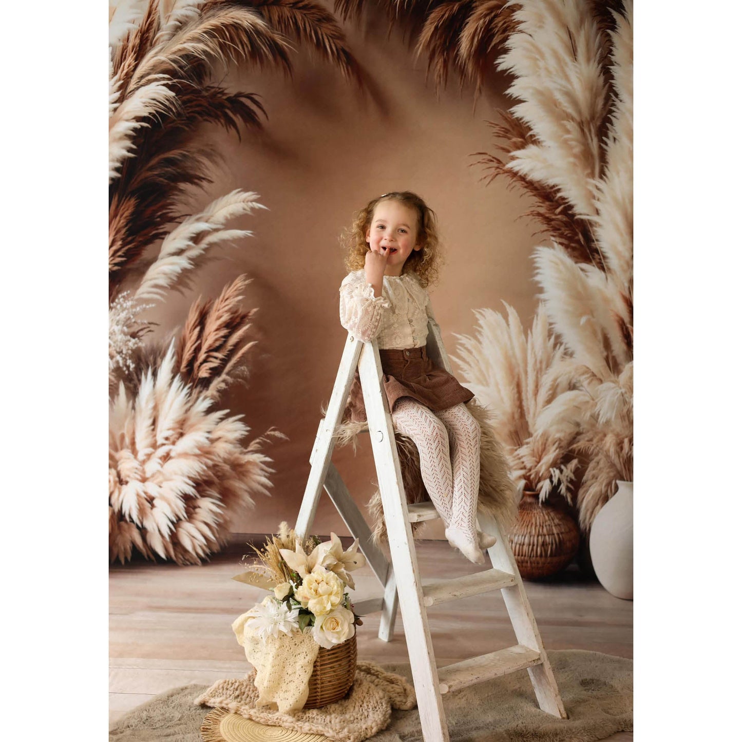 Young girl sitting on a ladder in front of pampas grass with a Sweep Boho Floral Arch Beige Backdrop in the background