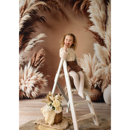 Young girl sitting on a ladder in front of pampas grass with a Sweep Boho Floral Arch Beige Backdrop in the background