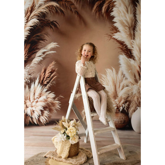 Young girl sitting on a ladder in front of pampas grass with a Sweep Boho Floral Arch Beige Backdrop in the background