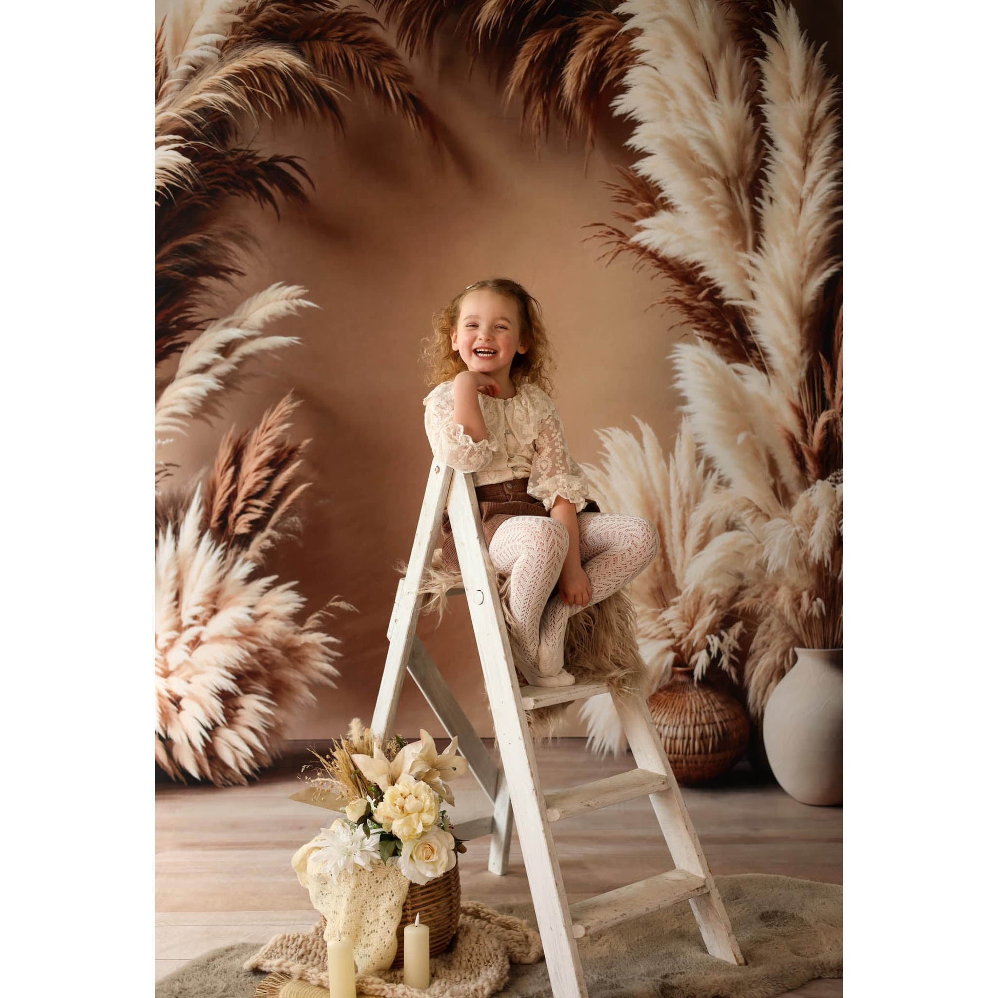 Young girl sitting on a ladder in front of pampas grass with a Sweep Boho Floral Arch Beige Backdrop in the background