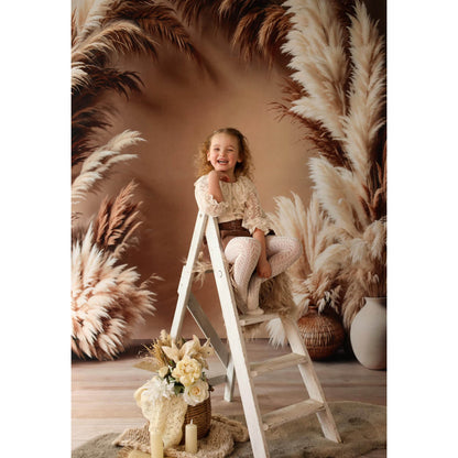 Young girl sitting on a ladder in front of pampas grass with a Sweep Boho Floral Arch Beige Backdrop in the background